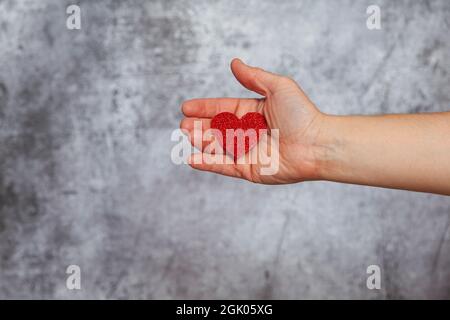 Die rechte Hand eines Mannes hält eine rote Glitzer-Herzform auf einem strukturierten grauen Hintergrund. Stockfoto