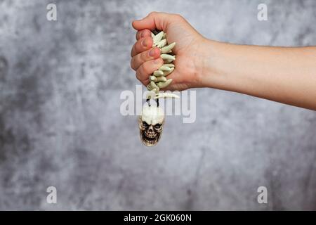 Die rechte Hand eines Mannes hält einen Anhänger mit Reißzähne, Knochen und einem kleinen Schädel auf einem strukturierten grauen Hintergrund. Stockfoto