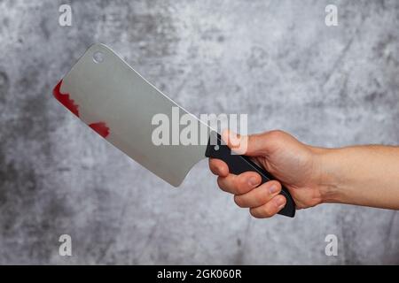 Die rechte Hand eines Mannes hält ein großes Schlachtermesser mit Blutflecken auf einem strukturierten grauen Hintergrund. Stockfoto