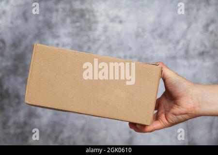 Die rechte Hand eines Mannes, der einen Karton hält oder liefert, der ein Paket sein kann, das auf einem strukturierten grauen Hintergrund geliefert wird. Stockfoto