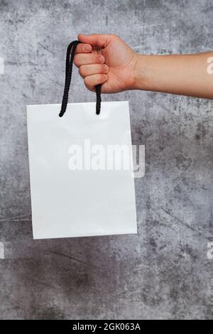 Die rechte Hand eines Mannes, der eine weiße Einkaufstasche mit schwarzen Seilgriffen auf einem strukturierten grauen Hintergrund hält. Stockfoto