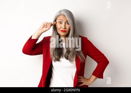 Verärgerte asiatische Geschäftsfrau schimpft jemand dumm, zeigt verrückte Geste, zeigt den Finger auf den Kopf und starrte auf die Kamera, weißer Hintergrund Stockfoto