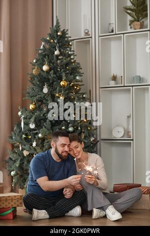 Fröhliches, junges kaukasisches Paar, das auf dem Boden gegen einen geschmückten Baum sitzt und zu Weihnachten Funker beleuchtet Stockfoto