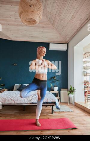 Ein fröhlicher Yogapraktiker in bequemer Sportkleidung steht in Baumstellung im hellen Raum Stockfoto