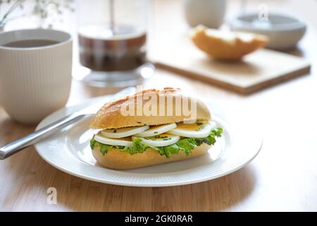 Sandwichrolle mit Eierscheiben, Salat und Schnittlauch auf einem weißen Teller garnieren und frischen Kaffee auf einem hellen Holzfrühstückstisch, ausgewählter Fokus, schmal Stockfoto