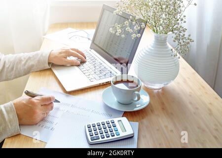 Hände arbeiten auf einem Schreibtisch im Home Office mit Laptop-Computer, Rechner, Papier, Kaffee und einem weißen Blumenstrauß, Business-Buchhaltung-Konzept, Kopie-SPAC Stockfoto