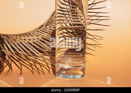 Goldenes Palmenblatt hinter transparentem Glas mit Wasser auf orangefarbenem Hintergrund. Optische Verzerrung durch Wasser. Nahaufnahme. Speicherplatz kopieren. Stockfoto