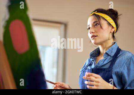 Porträt einer kreativen jungen Frau im Vorfeld, die ihre Arbeit während der Arbeit an abstrakter Malerei in einem gemütlichen Workshop betrachtet Stockfoto
