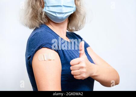 Geimpfte junge Frau mit Gipsschulter, weibliche Person in Maske Daumen nach Erhalt Coronavirus-Impfstoff. Konzept der Menschen COVID-19 vac Stockfoto