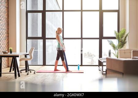 Seitenansicht einer fröhlichen, flinigen Frau, die mit einem Widerstandsband trainiert, Online-Videokurse auf einem Laptop ansieht, auf einer Yogamatte zu Hause in modernem Stil steht Stockfoto