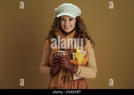 Hallo Herbst. Glücklich elegante 40 Jahre alte Hausfrau in Schal mit gelben Herbst Ahornblatt, Buch, Lederhandschuhe und Becher isoliert auf braunem Hintergrund. Stockfoto