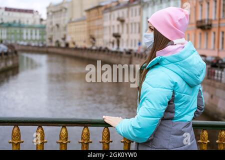 Eine junge Frau kaukasischer Ethnie geht in warmer Oberbekleidung und einer Schutzmaske durch die Stadt am Fluss, im Herbst geht sie während einer Pandemie um die Stadt Stockfoto