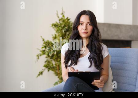 Eine junge Brünette füllt die Informationen aus dem Fragebogen aus. Die Frau schaut in die Kamera und zeichnet die Daten auf. Stockfoto