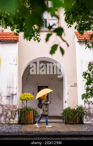 Frau mit gelbem Regenschirm läuft bei Regen auf der Stadtstraße. Weibliche touristische Spaziergänge in der europäischen Altstadt Kromeriz, Tschechische Republik Stockfoto