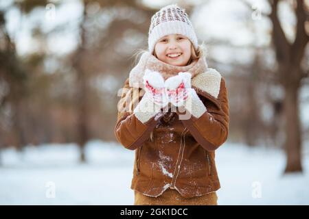 Lächelndes modernes Mädchen mit Fäustlingen in einem Strickhut und Schaffellmantel, das im Winter im Stadtpark spielt. Stockfoto