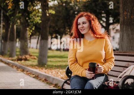 Porträt einer jungen rothaarigen Frau, die im Park auf der Bank mit einem Becher Kaffee sitzt. Lady trägt ein gelbes Sweatshirt und hält einen wiederverwendbaren Becher. Lifestyle Stockfoto