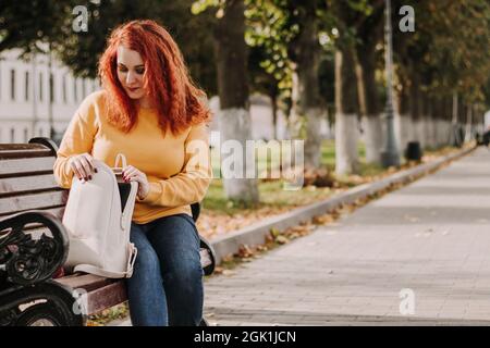 Die junge rothaarige Frau im gelben Pullover sitzt auf der Bank und sucht nach etwas in ihrer Tasche. Spazieren Sie an einem sonnigen Herbsttag. Stockfoto