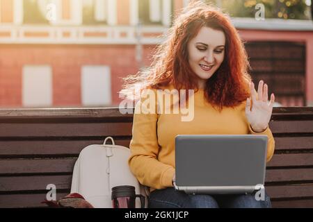 Rothaarige Frau in gelbem Pullover ist Video-Chat. Sie nutzt einen Laptop, um im Internet zu kommunizieren und zu arbeiten. Spaziergang am sonnigen Herbsttag. Stockfoto