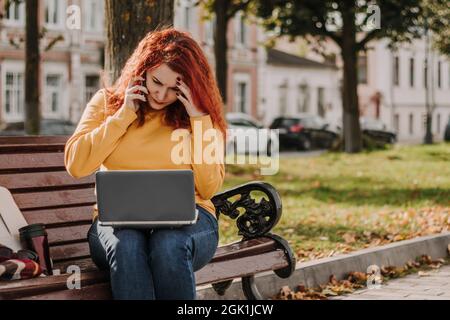 Die junge rothaarige Frau arbeitet im Park mit einem Laptop. Sie ist gestresst und löst Probleme. Moderne Online-Fernarbeit. Stockfoto