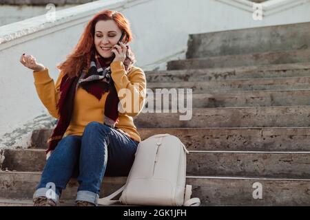 Junge rothaarige Frau, die auf einer Treppe sitzt und am Telefon spricht. Lady in gelb Pullover nutzt Smartphone an sonnigen Tag. Lifestyle, Kopierbereich. Stockfoto