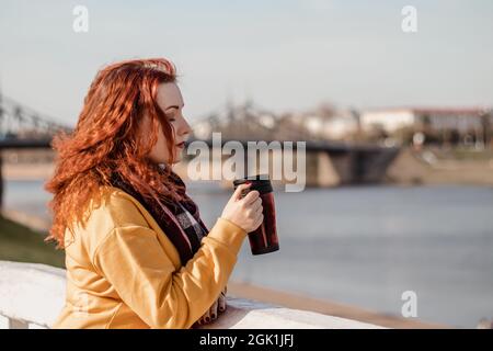 Junge rothaarige Frau in gelbem Pullover hält Thermobecher mit Kaffee in den Händen. Genießen Sie den Blick auf den Fluss am sonnigen Herbsttag. Umweltfreundlicher Lebensstil, Stockfoto