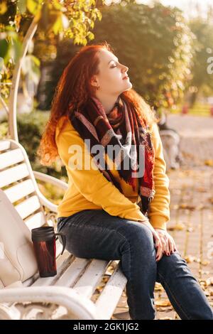 Junge rothaarige Frau, die auf der Bank im Park sitzt und den sonnigen Herbsttag genießt. Spazieren Sie bei Sonnenuntergang zwischen den gelben Bäumen. Stockfoto