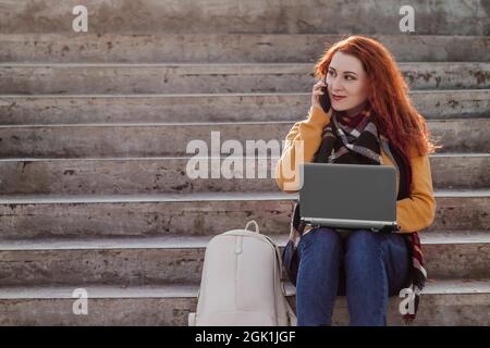 Die junge rothaarige Frau sitzt auf einer Treppe und kommuniziert über einen Laptop im Internet. Die moderne Geschäftsfrau arbeitet freiberuflich im Freien. Hipster mit Gadg Stockfoto