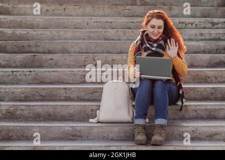 Die junge rothaarige Frau sitzt auf einer Treppe und kommuniziert über einen Laptop im Internet. Die moderne Geschäftsfrau arbeitet freiberuflich im Freien. Hipster mit Gadg Stockfoto