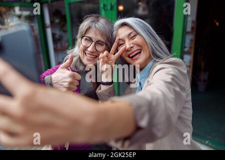 Attraktive reife asiatische Frau mit Freund nehmen Selfie gestikulieren mit Telefon auf der Straße der Stadt Stockfoto