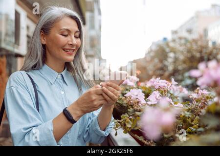 Lächelnde grauhaarige asiatische Frau benutzt Handy, das auf der Außenterrasse steht Stockfoto