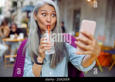 Lustige reife asiatische Frau trinkt Wasser nehmen Selfie mit Handy im Café Stockfoto