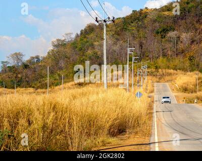 Einsames weißes Auto auf der Landstraße neben dem gelben Grasblumenfeld mit einer Reihe elektrischer Pole in der Nähe des Berges und blauem Himmel auf einer Sonne Stockfoto