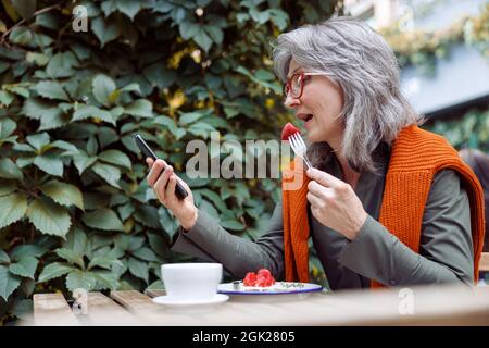 Reife Frau mit Brille isst Dessert und schaut auf Smartphone am Tisch im Freien Stockfoto