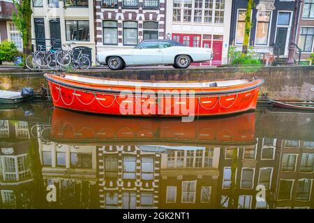 Amsterdam Holland - August 19 2017; Vintage Orange Farbe Grachtenboot neben mit weißen amerikanischen Oldtimer auf der Straße vor typica geparkt Stockfoto