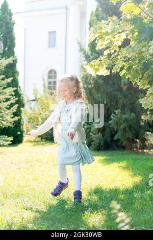 Kind wirbelt, tanzt auf der Wiese. Kleines Mädchen mit blondem Haar wirbelt in der Sonne. Lockiges Mädchen genießt das Leben. Frohe Kindheit. Stockfoto