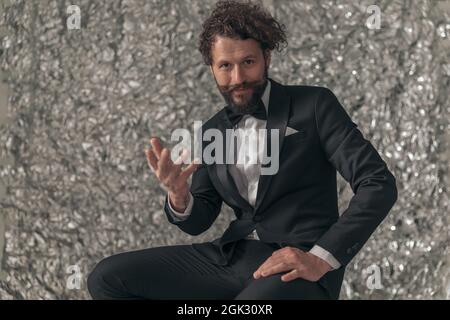 Porträt eines glücklichen, bärtigen, eleganten Mannes in schwarzem Smoking, der die Hand hochhält, gestikulierend und einladend, während er im Studio auf einem Kinfoil-Hintergrund posiert Stockfoto