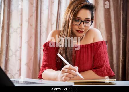 Lächelnde junge indische Frau korrigiert Dokument auf Tablet-Computer und Notizen mit digitalem Stift Stockfoto