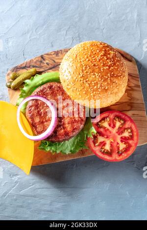 Hamburger Zutaten, oben geschossen auf blauem Hintergrund. Rinderfleisch, Cheddar-Käse, Zwiebeln, Tomaten, Salat und Sesambrotbrötchen, von oben geschossen Stockfoto