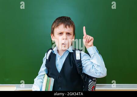 Lustige Schuljunge Grimassen in der Nähe der grünen Schultafel im Klassenzimmer. Grundschulkind mit Tasche und Büchern. Zurück zur Schule. Stockfoto