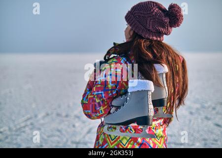 Eine Frau in einem Skianzug hält Schlittschuhe auf ihrer Schulter und blickt in die Ferne, nachdem sie auf einem gefrorenen See Skifahren konnte Stockfoto