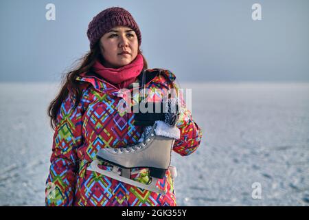 Eine Frau in einem Skianzug hält Schlittschuhe auf ihrer Schulter und blickt in die Ferne, nachdem sie auf einem gefrorenen See Skifahren konnte Stockfoto