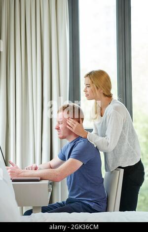 Hübsche junge Frau massiert Freund leiden unter schweren Kopfschmerzen aufgrund der Arbeit den ganzen Tag am Computer Stockfoto