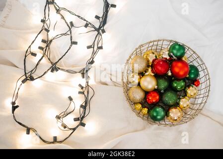 Rote, grüne und goldene Weihnachtskugeln liegen in einem Korb, neben einer glühenden Weihnachtsgirlande liegt auf einem weißen Tuch Stockfoto