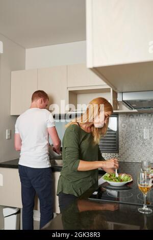 Junge Frau, die hausgemachten Salat mischt, als ihr Freund im Spülbecken Geschirr abspült Stockfoto