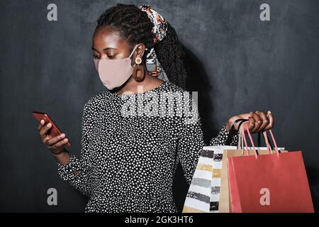 Afrikanische junge Frau in Maske mit ihrem Handy zum Online-Shopping vor dem schwarzen Hintergrund stehend Stockfoto