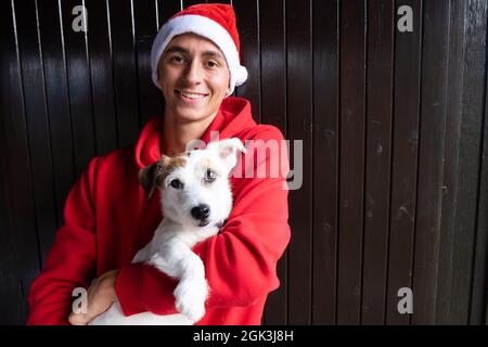 Weihnachtshund und junger Mann mit Weihnachtshut. Glückliche Momente, lächelndes Gesicht zu Weihnachten. Grüße und Freude mit Terrier Hund. Stockfoto