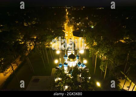 Luftaufnahme der beleuchteten Kirche zwischen Kiefern in der Nacht. Neue öffentliche leere weiße Kapelle mit goldenen und blauen Kuppeln im Freien von oben. Schöner religiöser, heiliger Ort für Ämter Stockfoto
