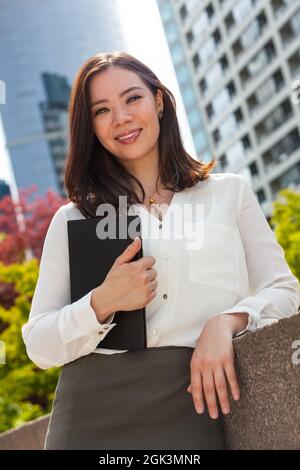 Porträt einer jungen asiatischen Frau oder eines Geschäftsmanns, die in einer Stadt lächelt und einen Ordner oder Tablet-Computer hält Stockfoto