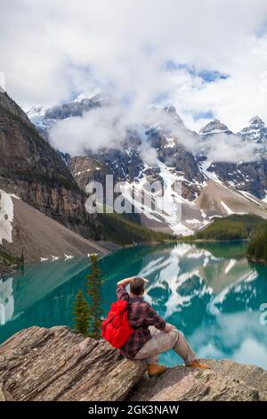 Wandermann mit rotem Rucksack auf einem Felsen mit Blick auf den Moraine Lake und Blick auf schneebedeckte Rocky Mountain-Gipfel, Banff National Park, Al Stockfoto