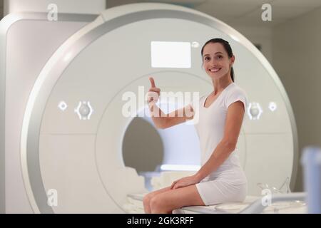 Junge Frau, die auf einer Magnetresonanztomographie-Maschine sitzt und den Daumen nach oben zeigt Stockfoto